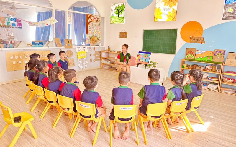 Các bé ngồi vòng tròn nghe cô trong lớp học sáng sủa